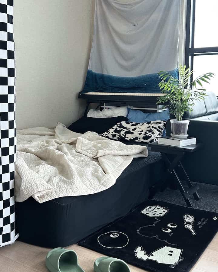 Black and white toned studio bedroom featuring a black bed with white bedding, pillows, checkered curtain, rug, and a green plant creating a cozy atmosphere