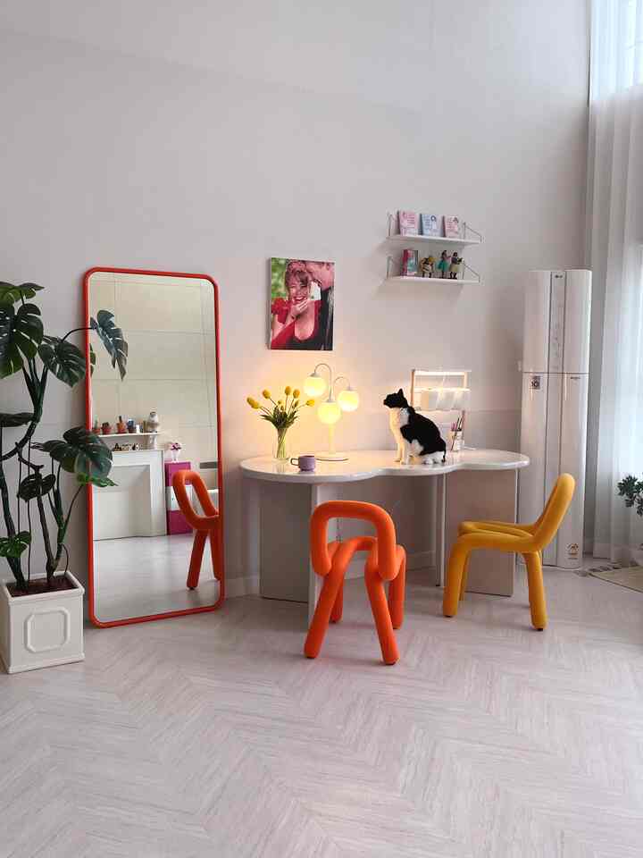 明るい色調のミッドセンチュリーモダンなリビング、オレンジと黄色の椅子、白い曲線のダイニングテーブルに座る黒白の猫とオレンジ色の全身鏡が配置された印象的な空間