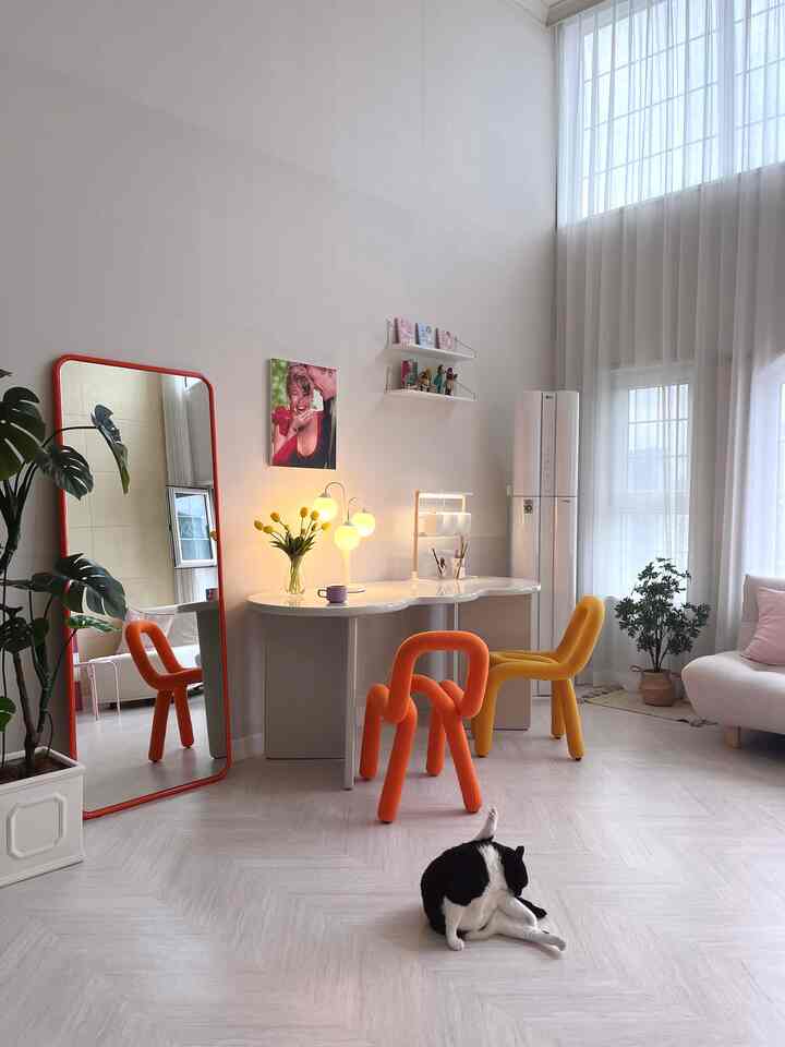 明るい白い壁と高い天井のリビング、曲線的なオレンジ色と黄色の椅子、白いデスクが配置され、床で毛づくろいをする猫がいる印象的な空間