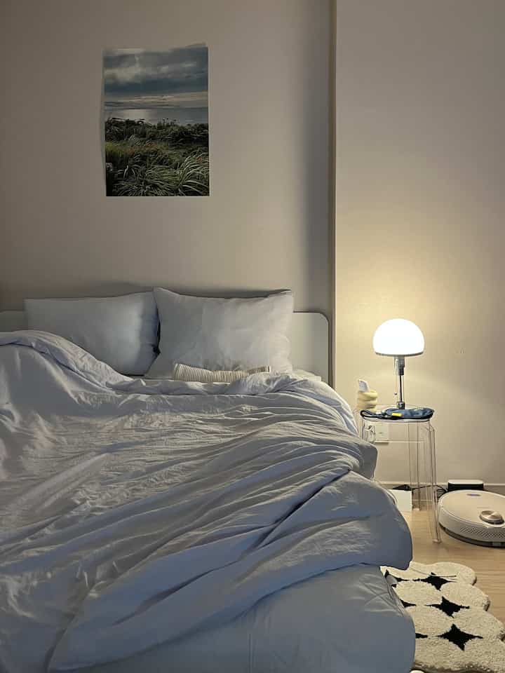 Pastel-toned studio bedroom in white featuring clean bedding and acrylic side table with warm lighting