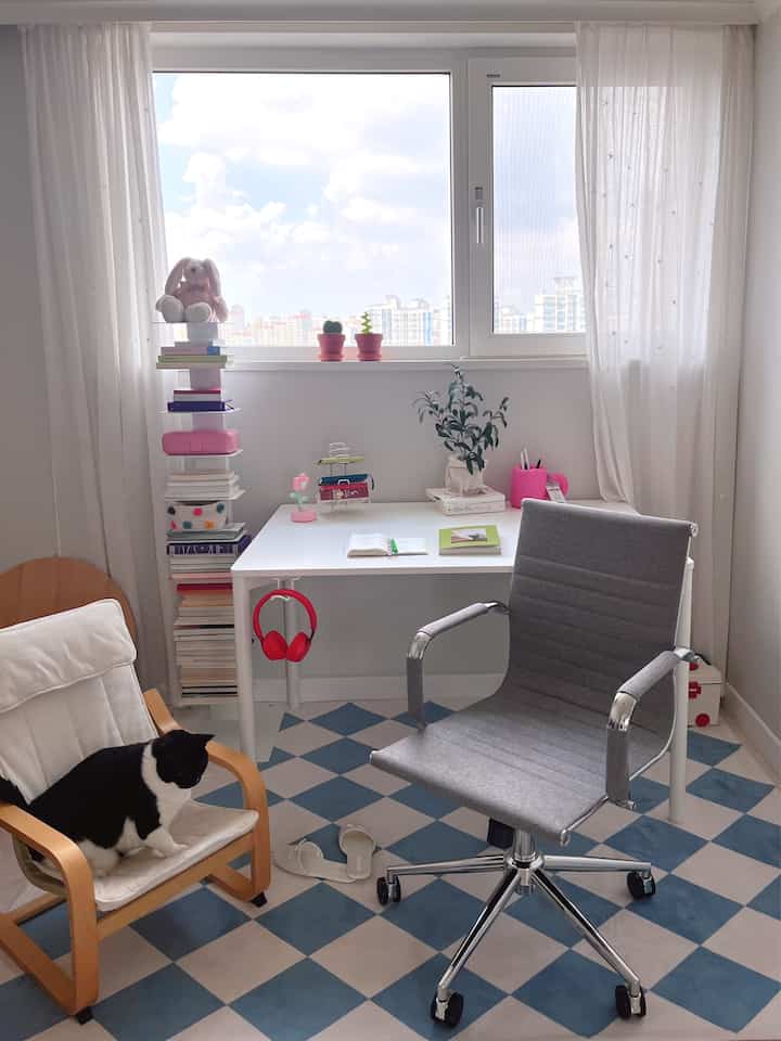 明るい白と青のチェック柄の床がある書斎、グレーのオフィスチェア、木製のアームチェア、猫がいる居心地の良い作業空間