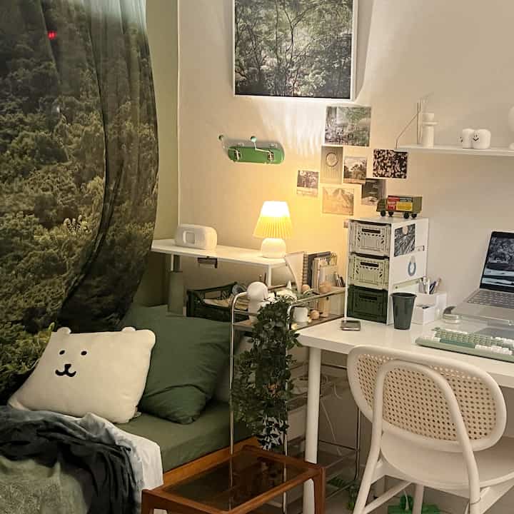 A cozy small studio room with natural tones, featuring a forest fabric poster, green cushions, and a clean white desk with warm lighting creating a restful home office.