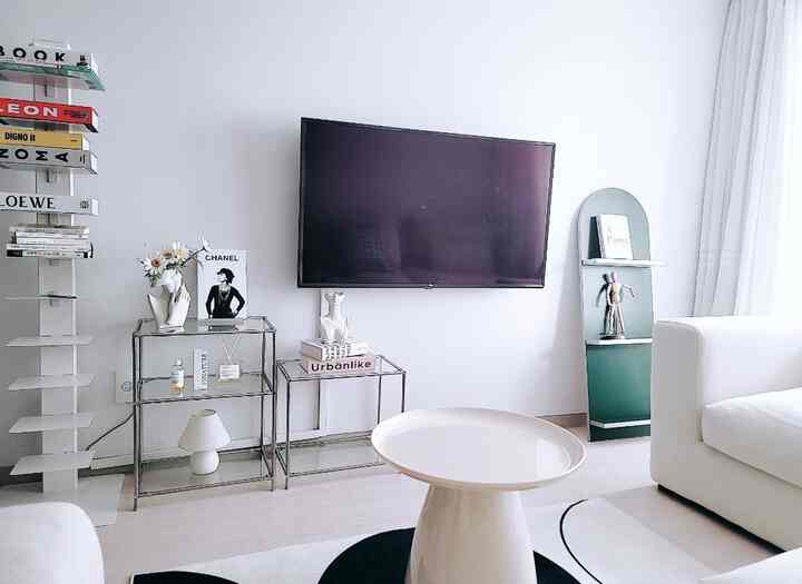 明るい白を基調としたリビングルーム、壁掛けテレビ、白いソファ、丸いコーヒーテーブル、本棚が配置されたシンプルなインテリア