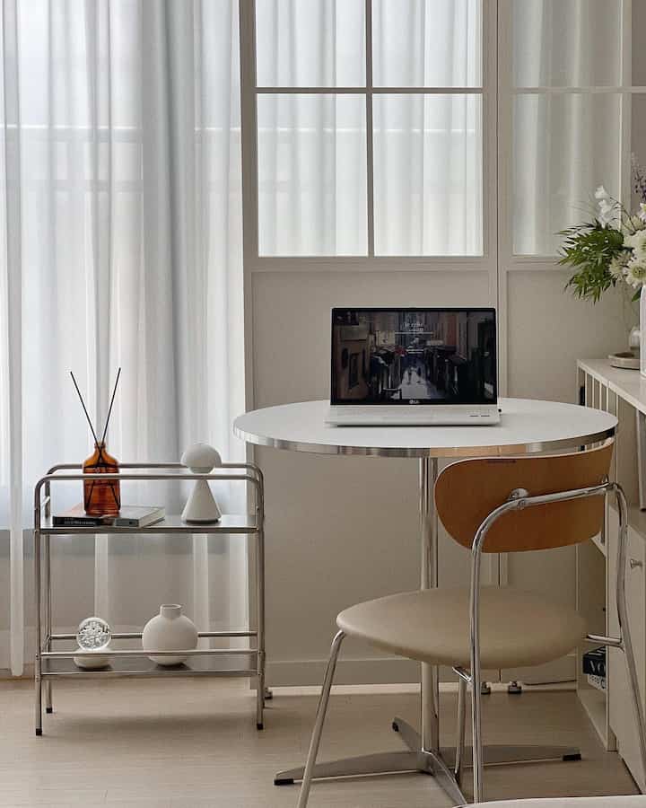 A white-toned compact dining room featuring a round table with chairs, glass room divider, diffuser, and bookshelf in a simple and clean setting