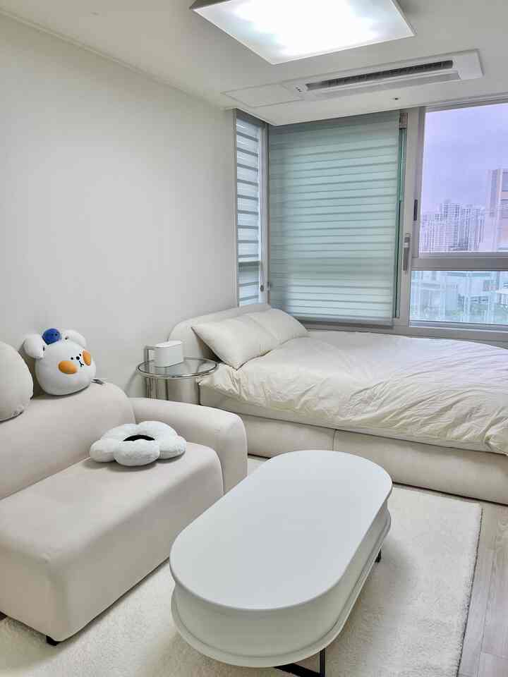 White-toned studio apartment combining living room and bedroom with two-seater sofa, bed, and coffee table in a clean modern space