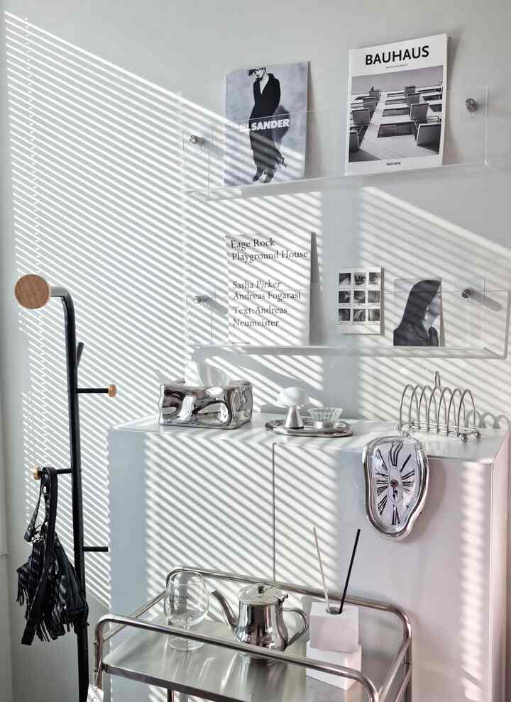 白とシルバーを基調としたワンルーム、ミッドセンチュリーモダンのコートラックとトロリー、デザイン時計と壁棚が配置された印象的なインテリア