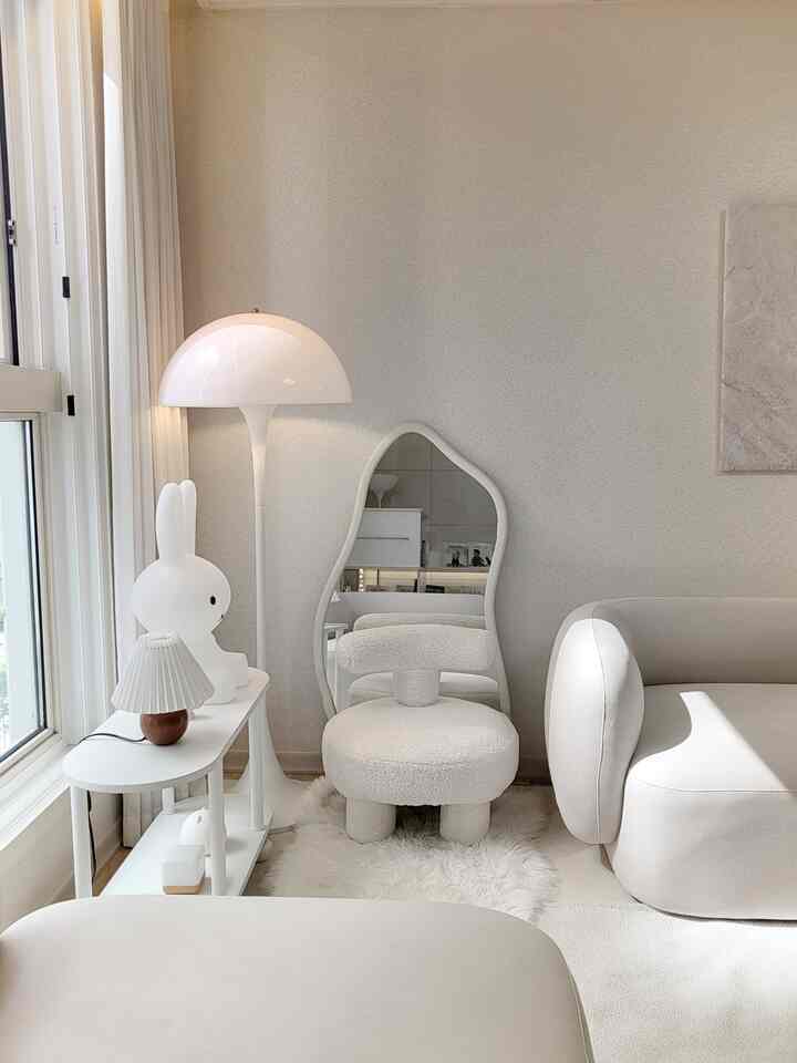 柔らかいホワイトトーンのリビング、曲線的なアームチェアとフロアランプ、不規則な形の鏡が配置された自然でシンプルな印象的な空間