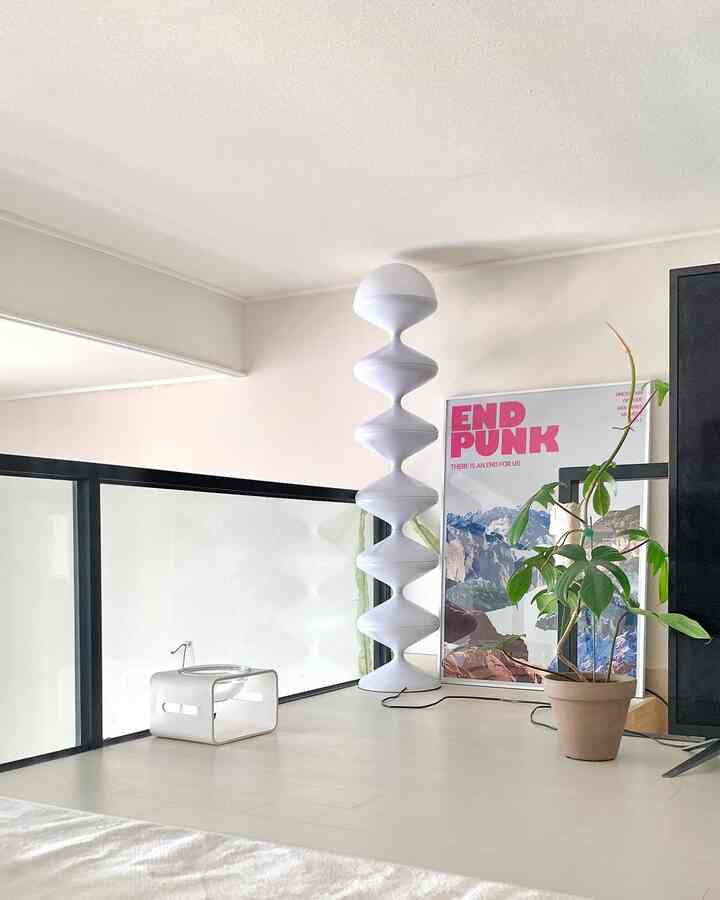 白と黒のトーンの複層ワンルームリビング、大きな彫刻的なフロアランプとピンクの文字ポスター、中型の観葉植物が配置されたミニマルなインテリア空間
