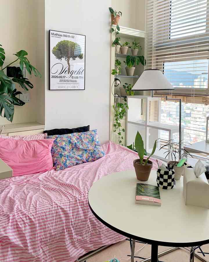 ベージュ調のワンルーム寝室、ピンクの寝具と植物、黒い額縁のポスターが配置された雰囲気のあるインテリア