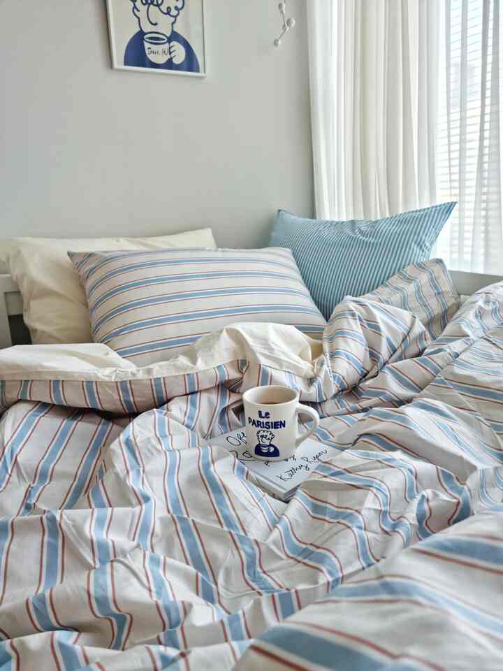 A cozy bedroom with white and blue tones, featuring striped bedding set and a mug on the bed