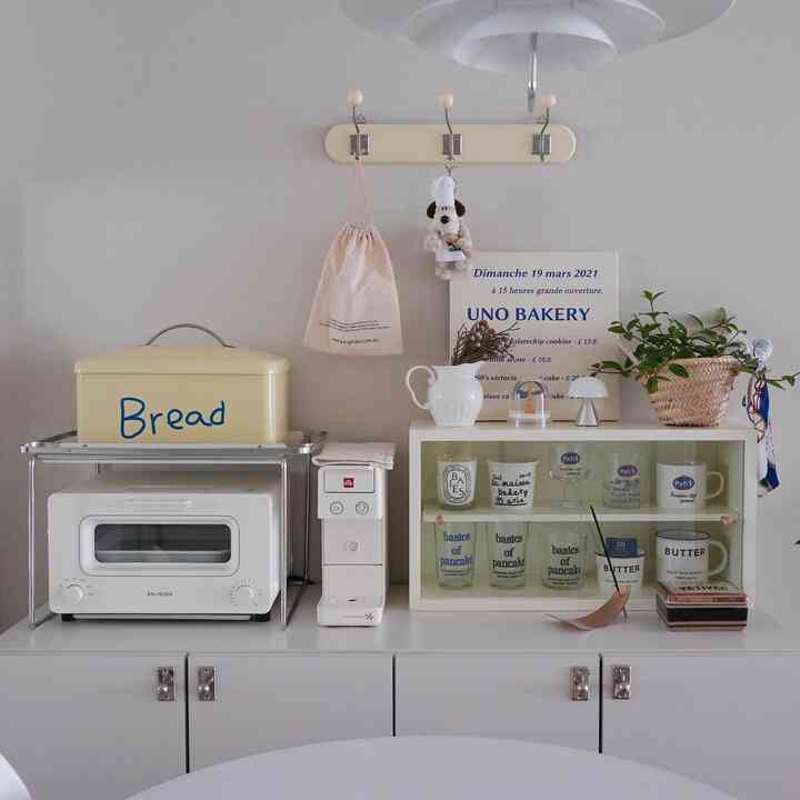 明るいホワイトトーンのキッチンおうちカフェゾーン、カプセルコーヒーマシンとパンボックス、植物の小物が配置された温かみのある空間