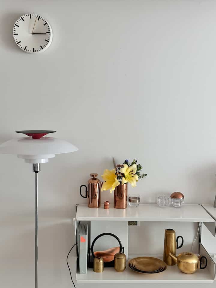 白い壁と棚のあるリビングルーム、金色と銅色の装飾品とフロアランプおよび壁掛け時計が配置されたすっきりとしたインテリア