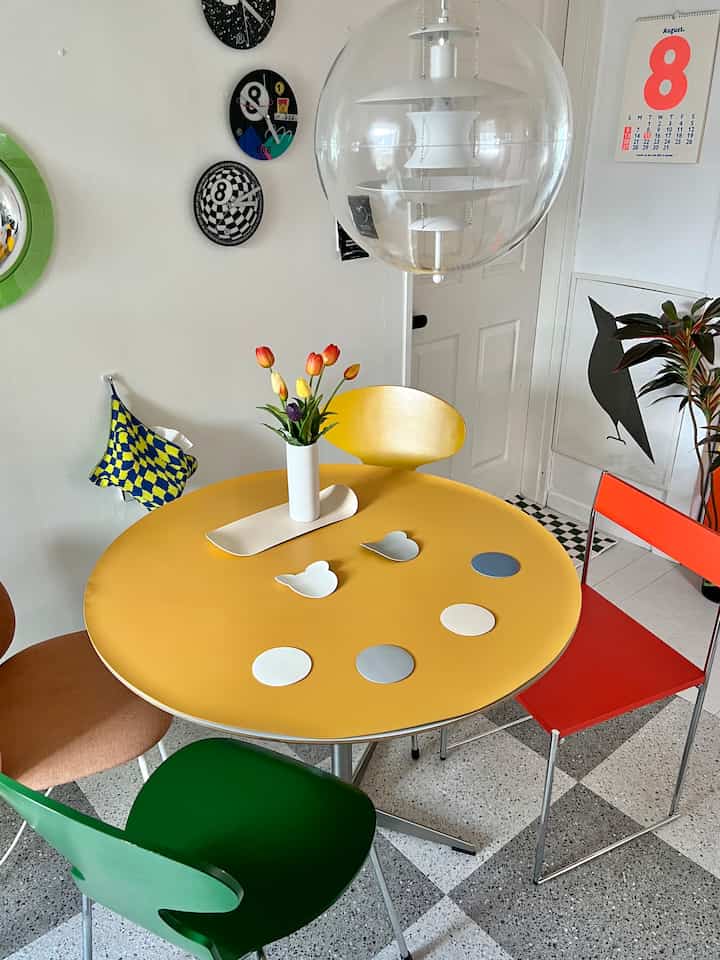 Bright yellow dining room featuring colorful chairs, a transparent pendant light, and wall clocks creating a cozy setting