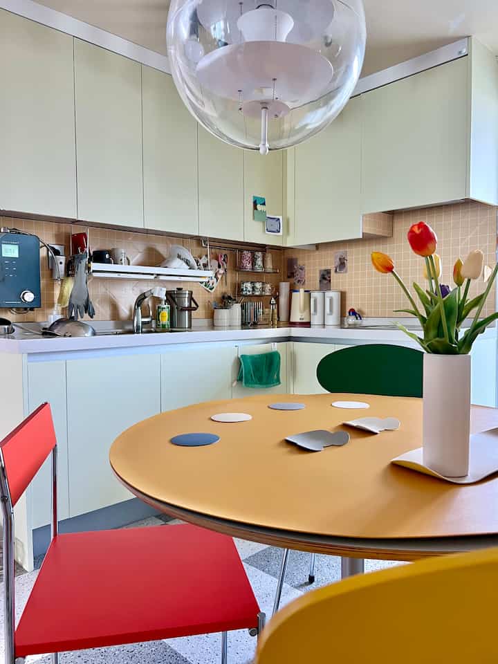 Pastel-toned kitchen featuring a yellow dining table, red chairs, and a tulip vase, creating a warm modern atmosphere