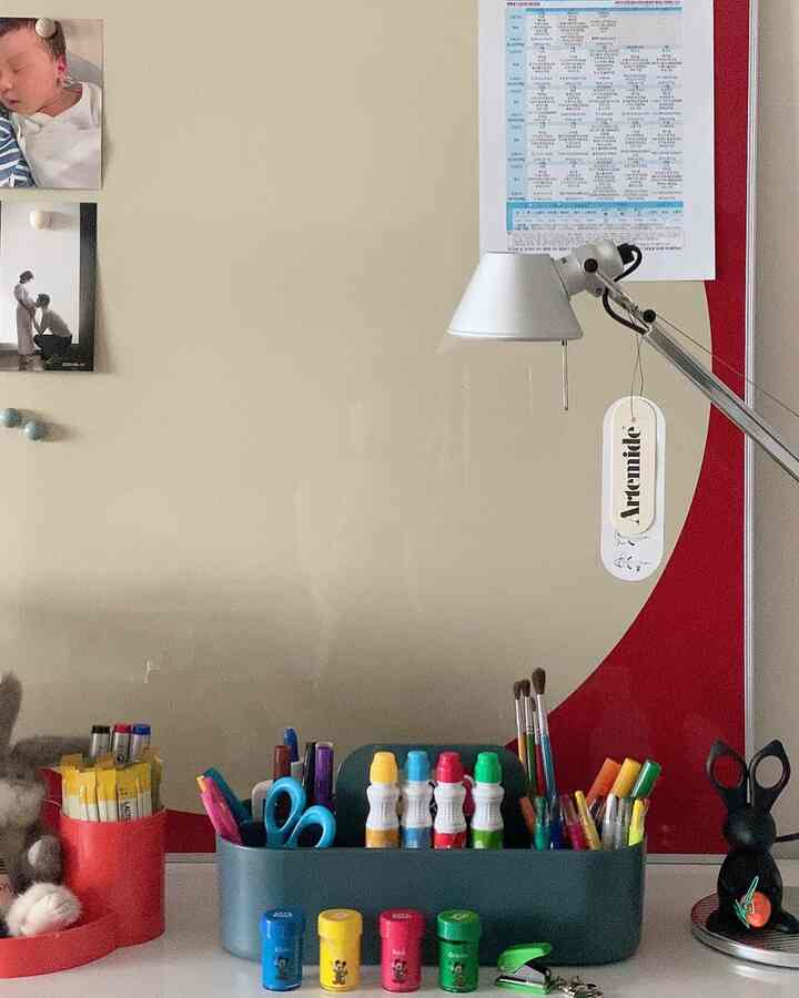 Simple kids' room with a red accent wall, baby photos on the wall, a desk filled with stationery, and a table lamp