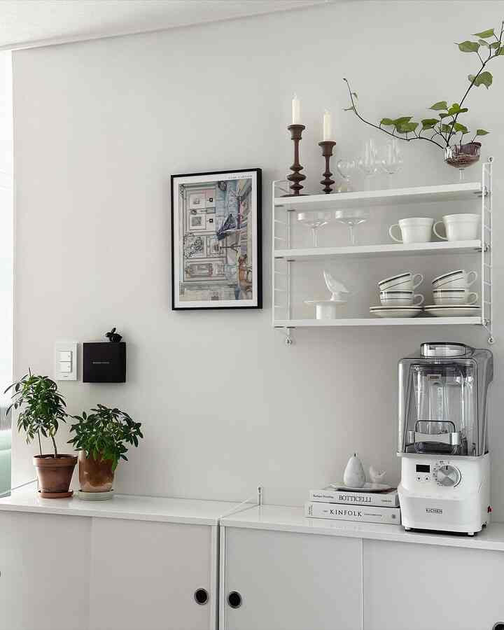 A modern kitchen space with white and natural tones featuring a wall shelf, plants, and a blender arranged neatly
