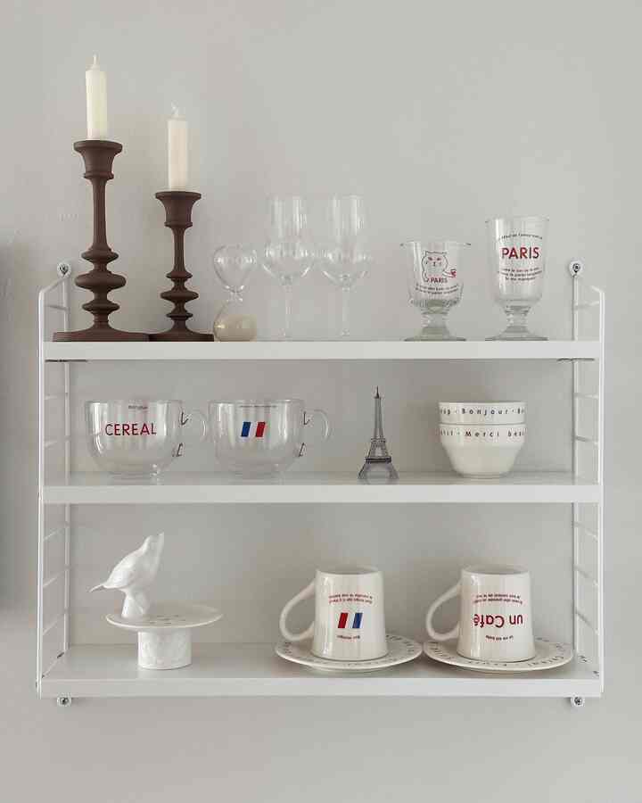 White-toned wall shelf with brown candlesticks, clear glassware, and mugs neatly arranged, creating a cozy home cafe vibe