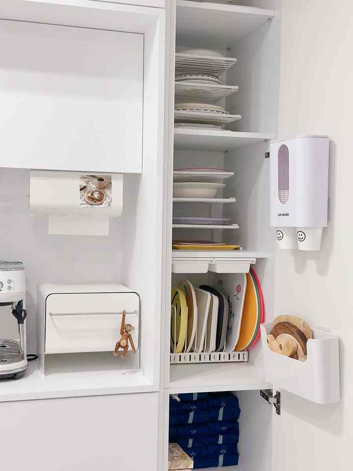 White-toned kitchen featuring a slim tall cabinet neatly storing plates, trays, and paper cups for a clean home cafe setup