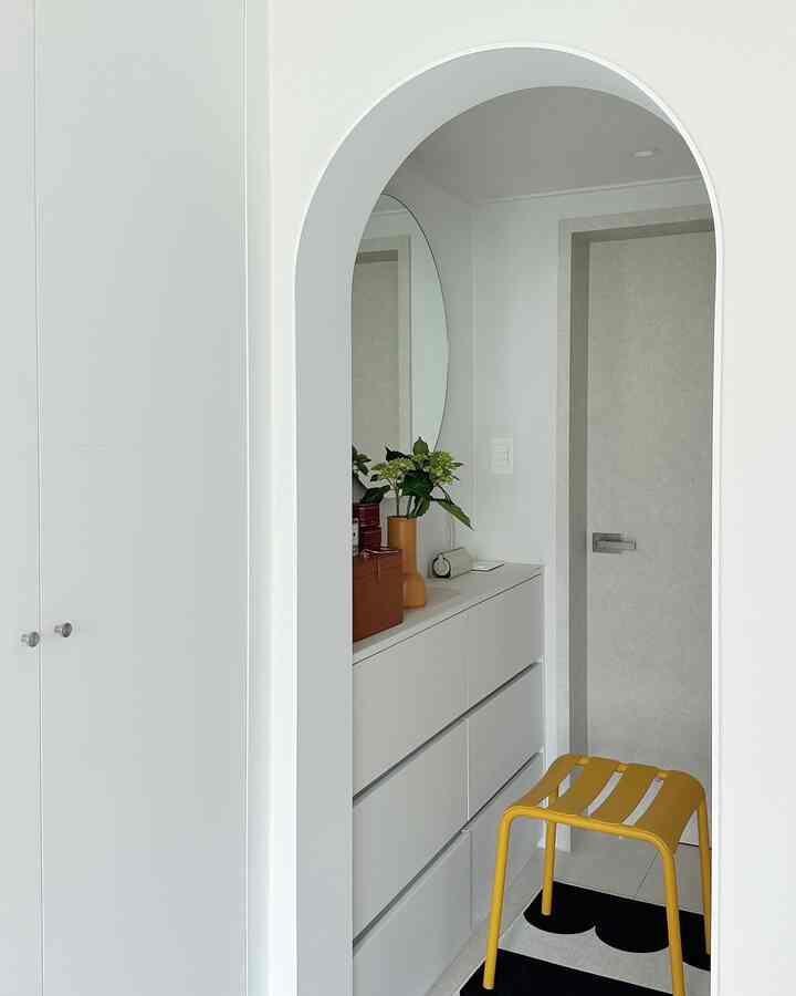 白を基調とした玄関、収納棚と丸い鏡が配置され、黄色のスツールがアクセントのシンプルな空間