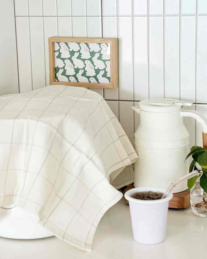 白と木目調が調和するキッチン空間、テーブルクロスやウサギ柄の額縁、電気ポットとカップが配置された清潔でミニマルな雰囲気のインテリア