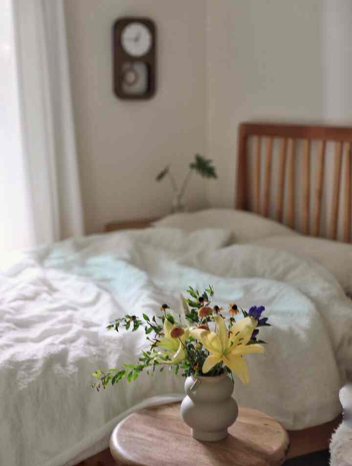 Natural wood-tone bedroom with wooden framed bed and white bedding, featuring a vase with flowers on a round stool in foreground center