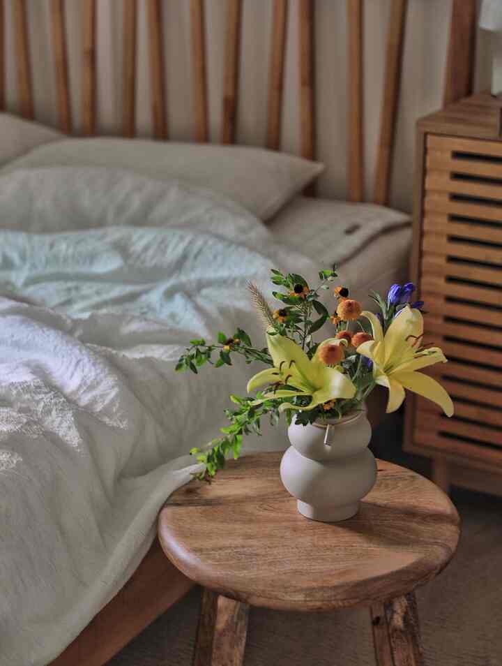 ナチュラルな寝室、白い寝具と木目調のスツールに淡い黄色のユリの花瓶が置かれた雰囲気のあるインテリア