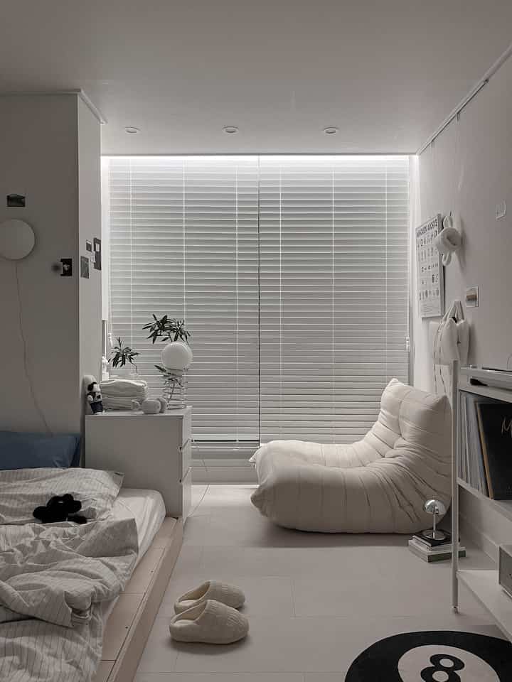 Simple modern bedroom in white and beige tones featuring a single sofa, bed, and large wooden blinds creating a clean and compact space