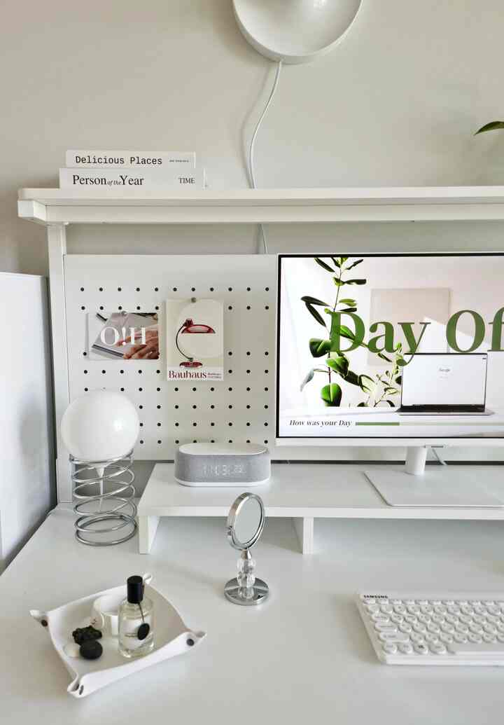 White-toned home office space featuring a desk with monitor, alarm clock, small mirror, and table lamp arranged neatly for a simple atmosphere