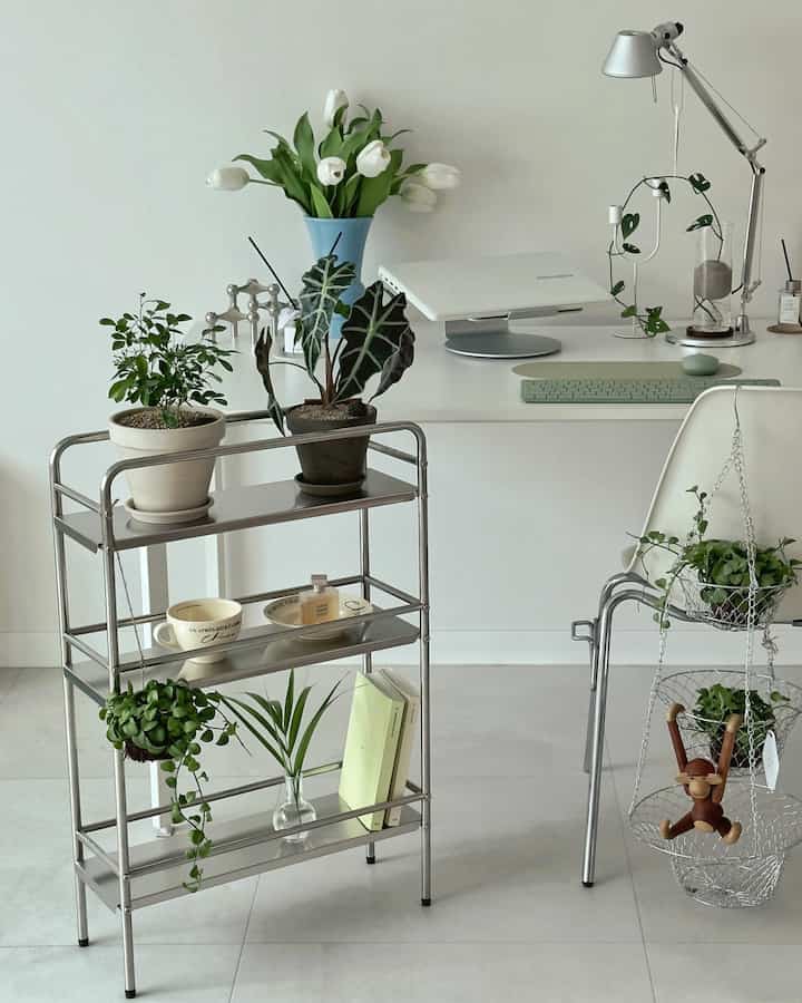 White and green toned home office space featuring metal shelving and desk adorned with various plants in a modern interior