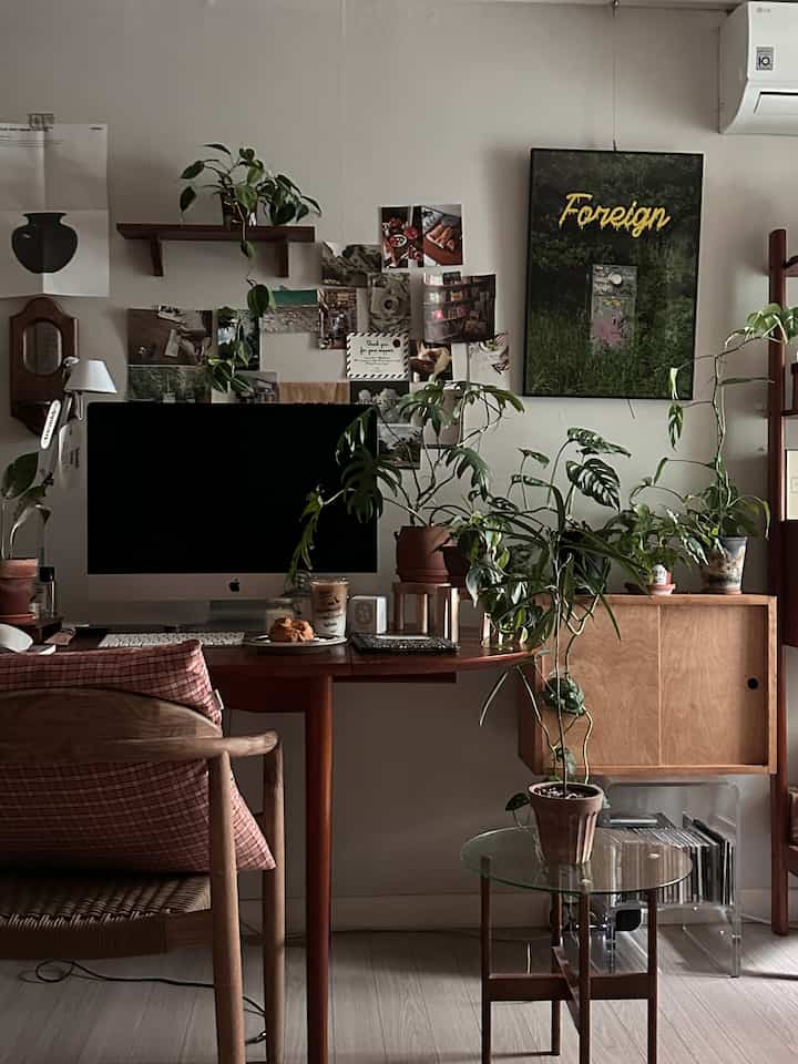 Natural-toned bedroom home office with white walls and colorful wall art, featuring wooden furniture and abundant plants creating a cozy atmosphere