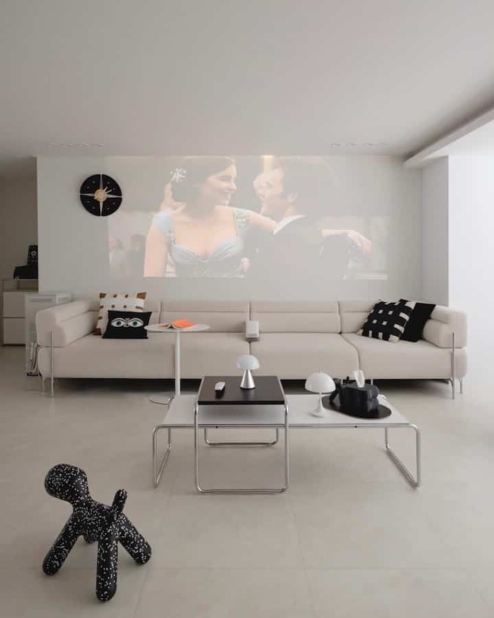 Beige-toned living room featuring a beige modular sofa, black and white coffee tables, and a home cinema projector display on the wall with simple modern decor