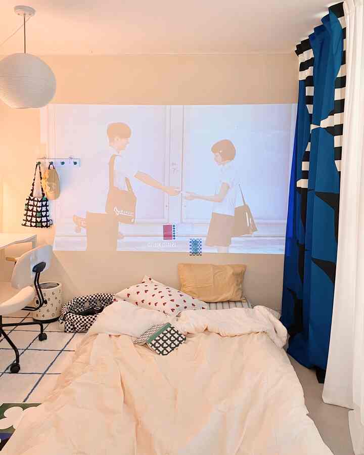 Beige-toned bedroom featuring a bed with pillows and comforter, blue patterned curtains, and a large projector screen on the wall creating a cozy atmosphere