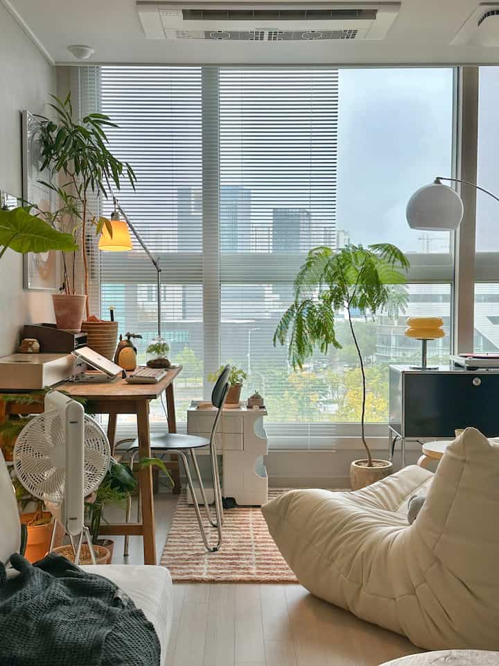 明るいベージュと白のワンルーム、木の植物とデスク、ソファが配置された自然な雰囲気のあるインテリア