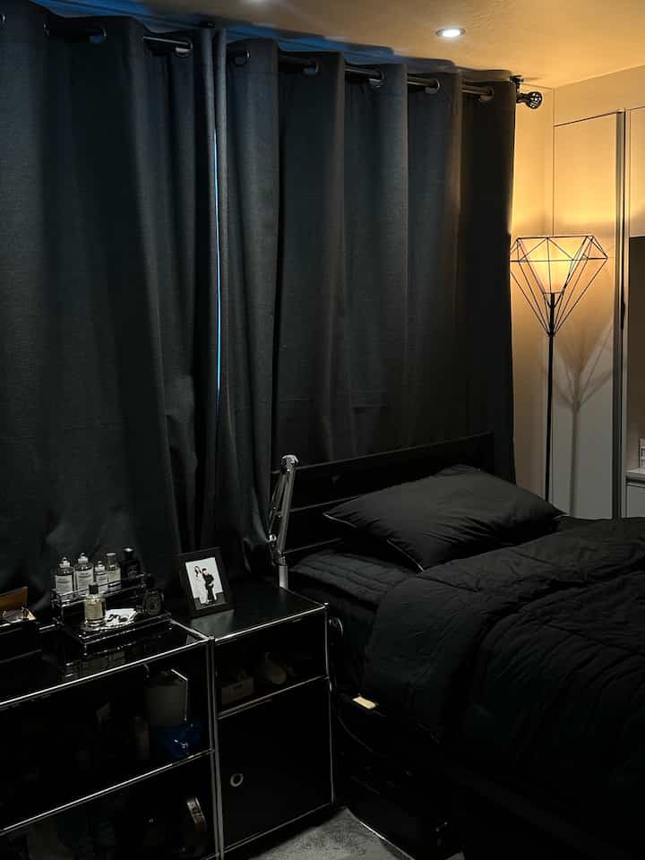 Dark black tone bedroom featuring black bed frame, black bedding, nightstand with accessories, blackout curtains, and a stylish floor lamp creating a modern calm atmosphere