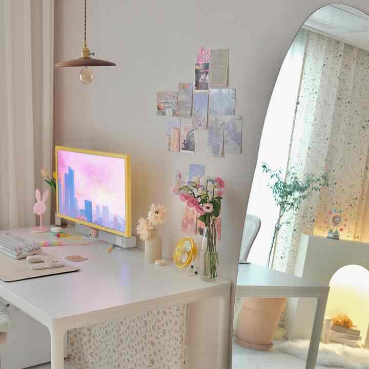 パステルトーンとホワイトを基調とした小さな部屋、中央に白いデスクと大型の鏡が配置され、壁にはポストカードのインテリアが並ぶ印象的な空間