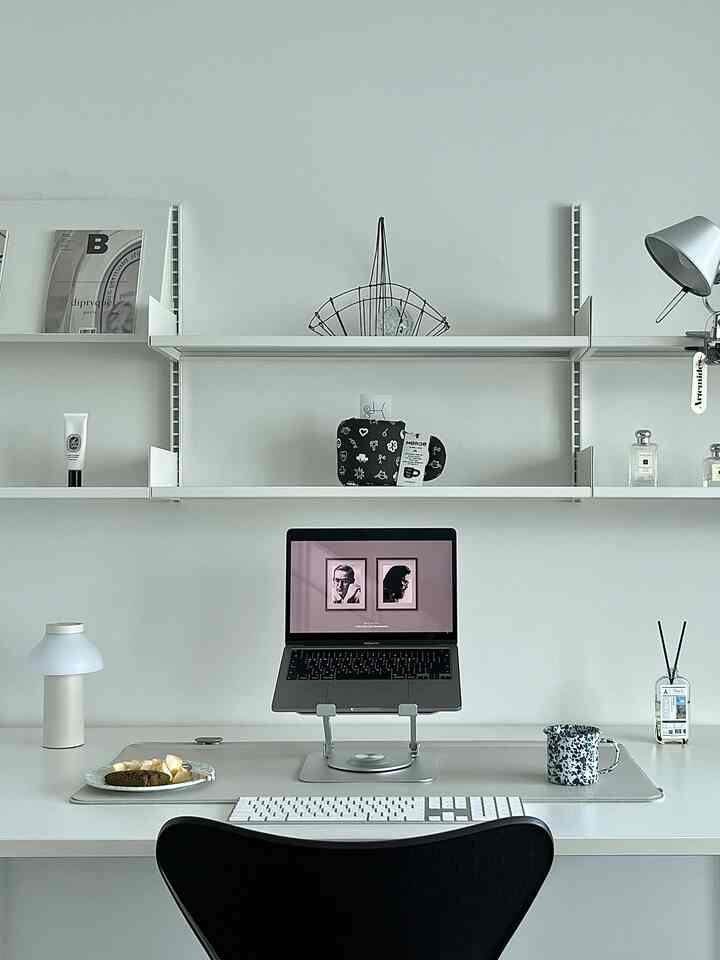 White-toned home office featuring a modern desk, laptop stand, and decorative objets on shelves for a clean workspace