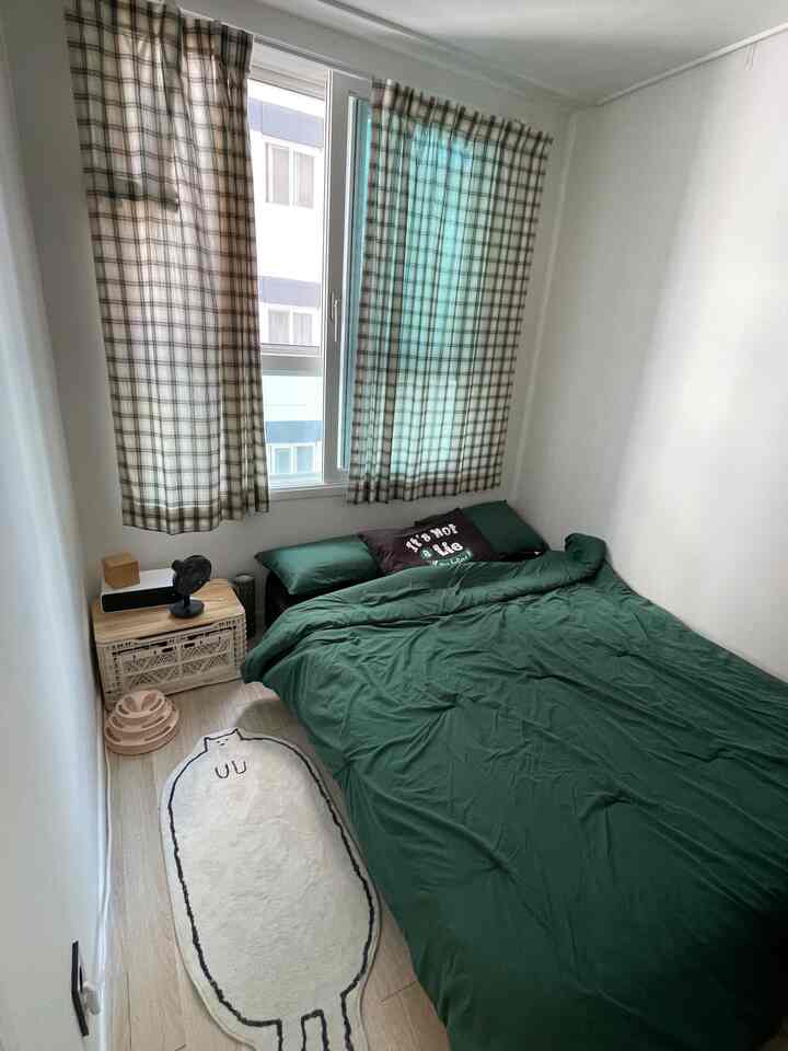 白い壁と床に緑の寝具、チェック柄のカーテンと猫形のラグが配置された狭くミニマルな寝室のインテリア