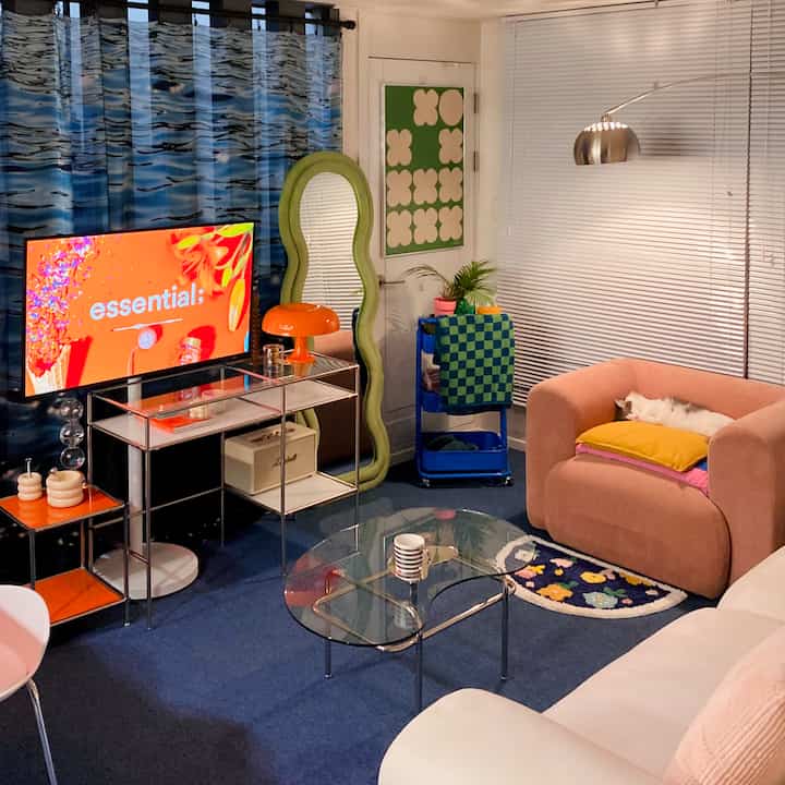 A mid-century modern living room with blue carpet and white walls, featuring a pink armchair with a white cat, an irregular green-framed full-length mirror, and a round glass coffee table in a colorful setting