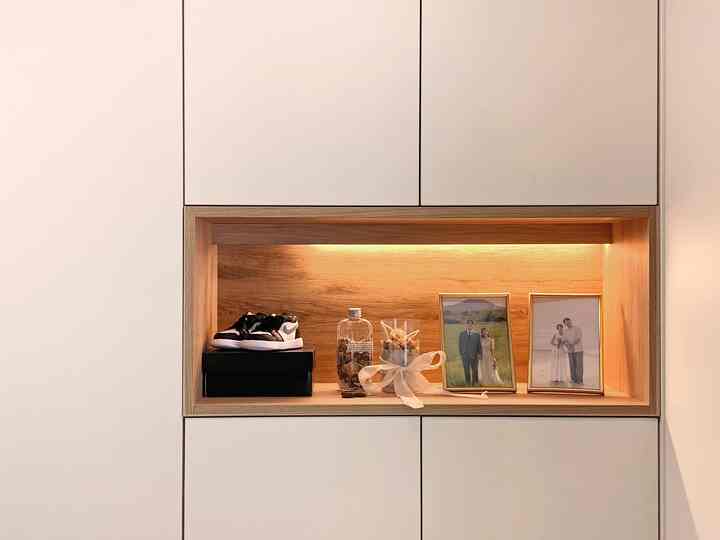 Natural wood-tone lit niche in shoe cabinet with picture frames, shoes, and objets, showcasing a simple modern entrance interior