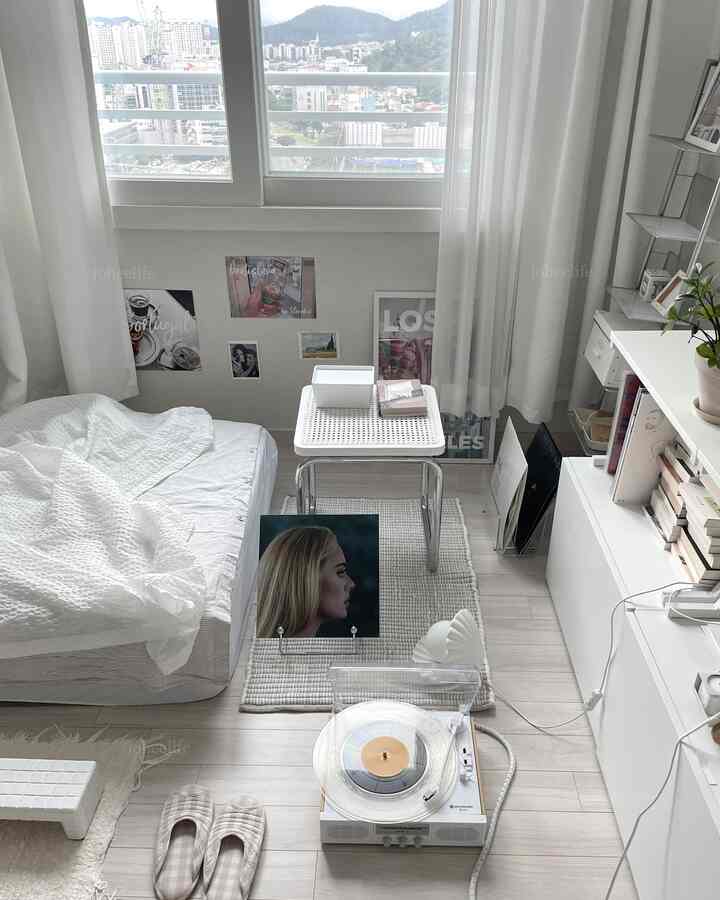 White-toned studio apartment living space featuring LP turntable, low bed, shelving, and curtains in a minimalist setup