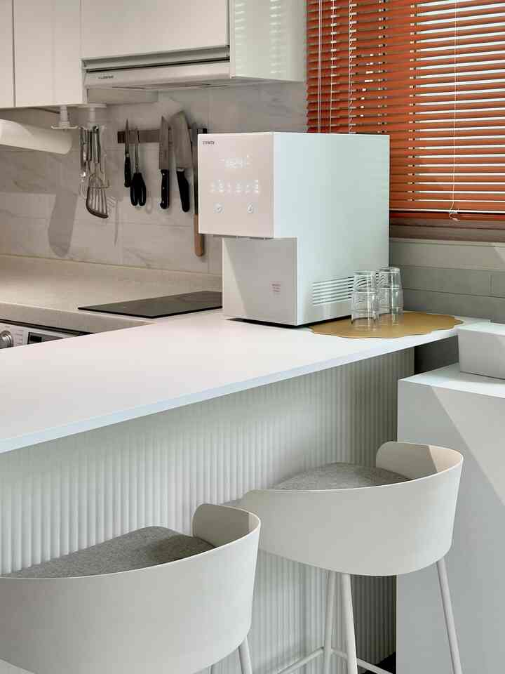白を基調としたモダンなキッチン空間で、バースツールとキッチンアイランドが配置され、狭い隙間にウォーターピューリファイアが置かれている印象的なインテリア。