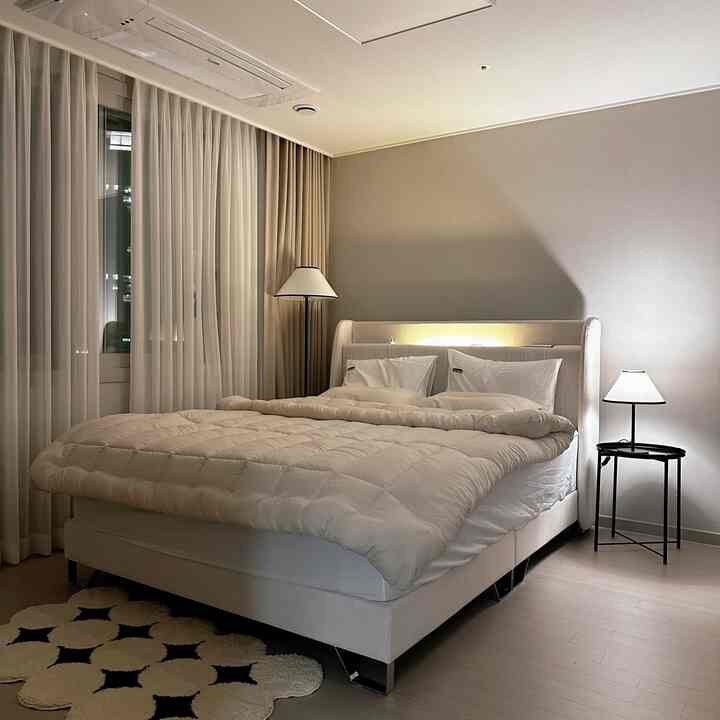 A cozy master bedroom in beige and black tones featuring a bed, nightstand, and standing lamps creating a warm modern atmosphere