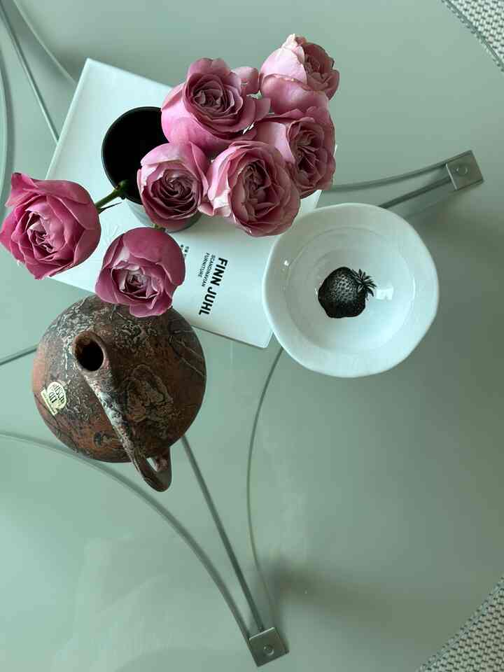 Minimal living room featuring transparent glass coffee table with pink rose vase, brown objet, and white plate with strawberry illustration