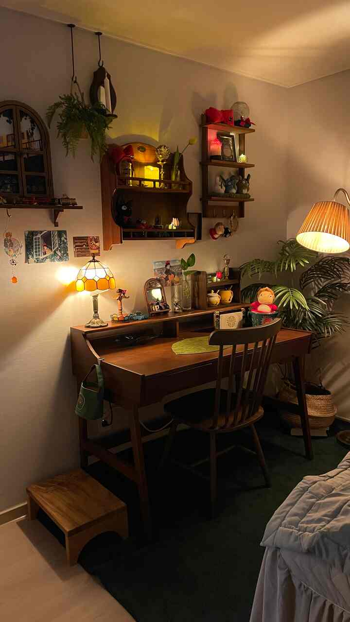 暖かい照明が灯る木製デスクと椅子のある居心地の良い部屋、ジブリのぬいぐるみと緑の植物が配置されている雰囲気のあるインテリア