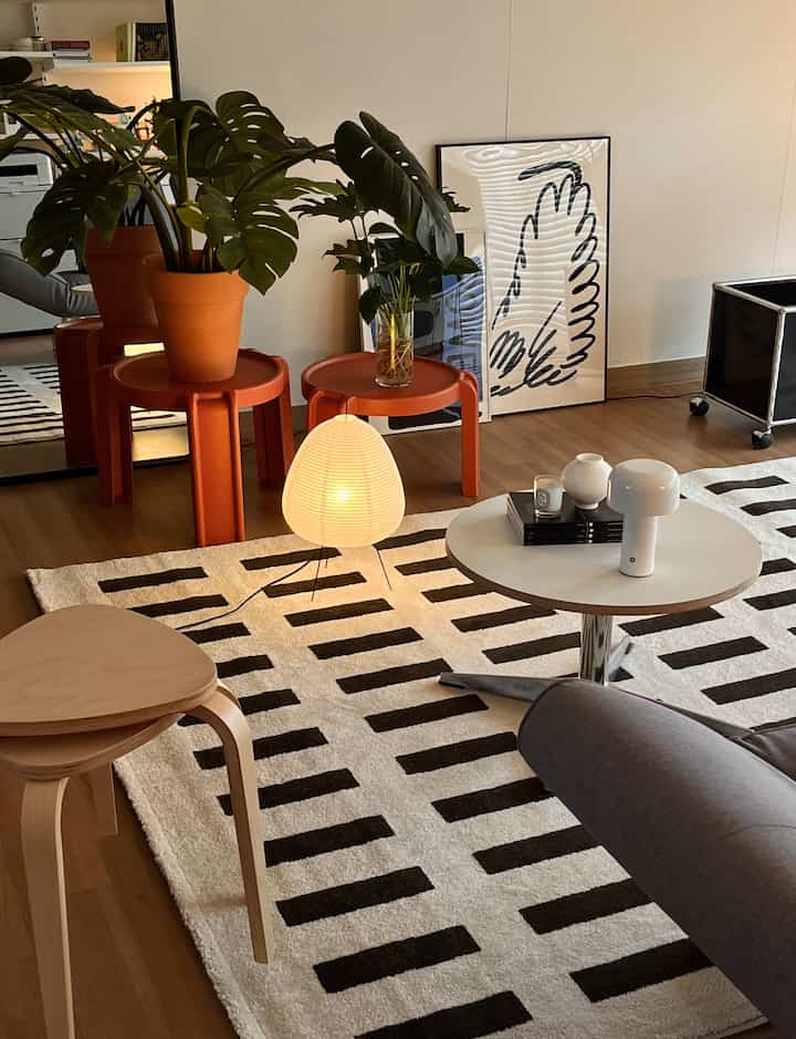 A modern living room with light wood flooring, white and orange side tables, and green plants arranged stylishly