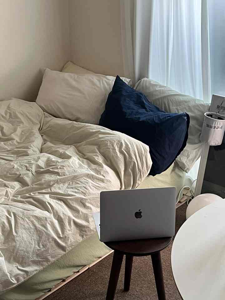 アイボリー色調の寝室、枕と布団が置かれたベッドとその前にあるダークウッドのスツールが配された、ナチュラルな雰囲気のインテリア