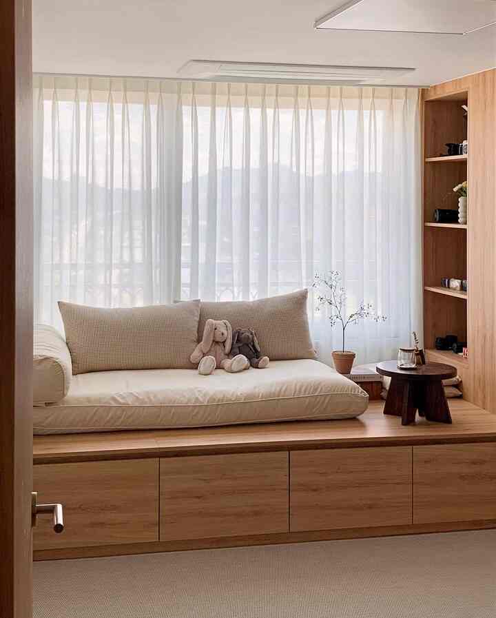 Bright beige cushions and stuffed toys on wood tone platform with large window in a minimal bedroom space
