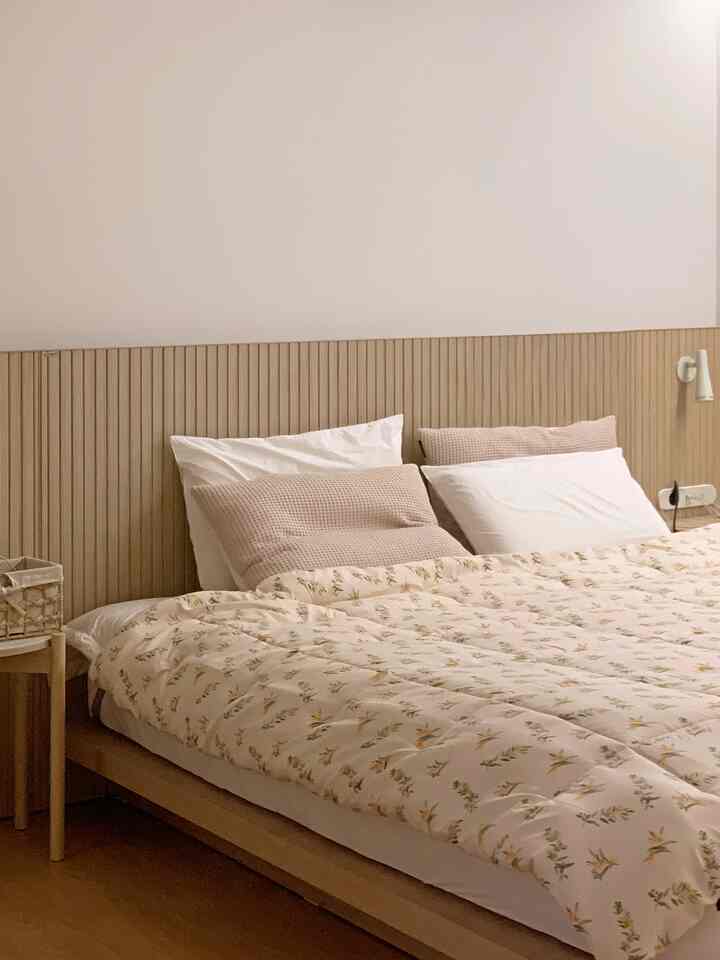 A natural wood tone beige bedroom featuring a hotel-like wood bed frame, pillows, and a nightstand in a minimal and warm space