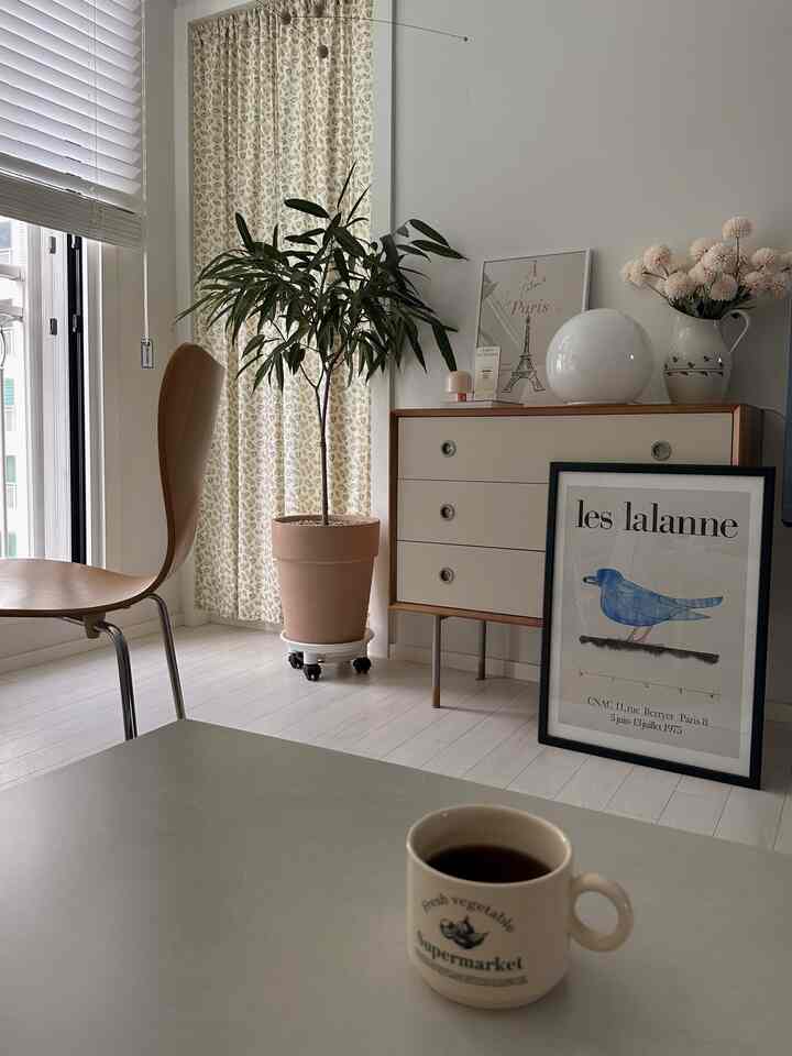 Natural toned living room featuring wood tone chair and dresser, plant and beige curtains creating a simple and harmonious space
