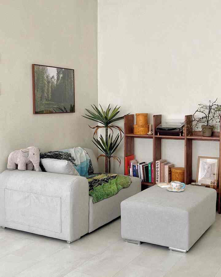 淡いベージュとグレーの色調のリビング、2人用のグレーソファとスツール、木製の本棚と植物が調和した雰囲気のあるインテリア